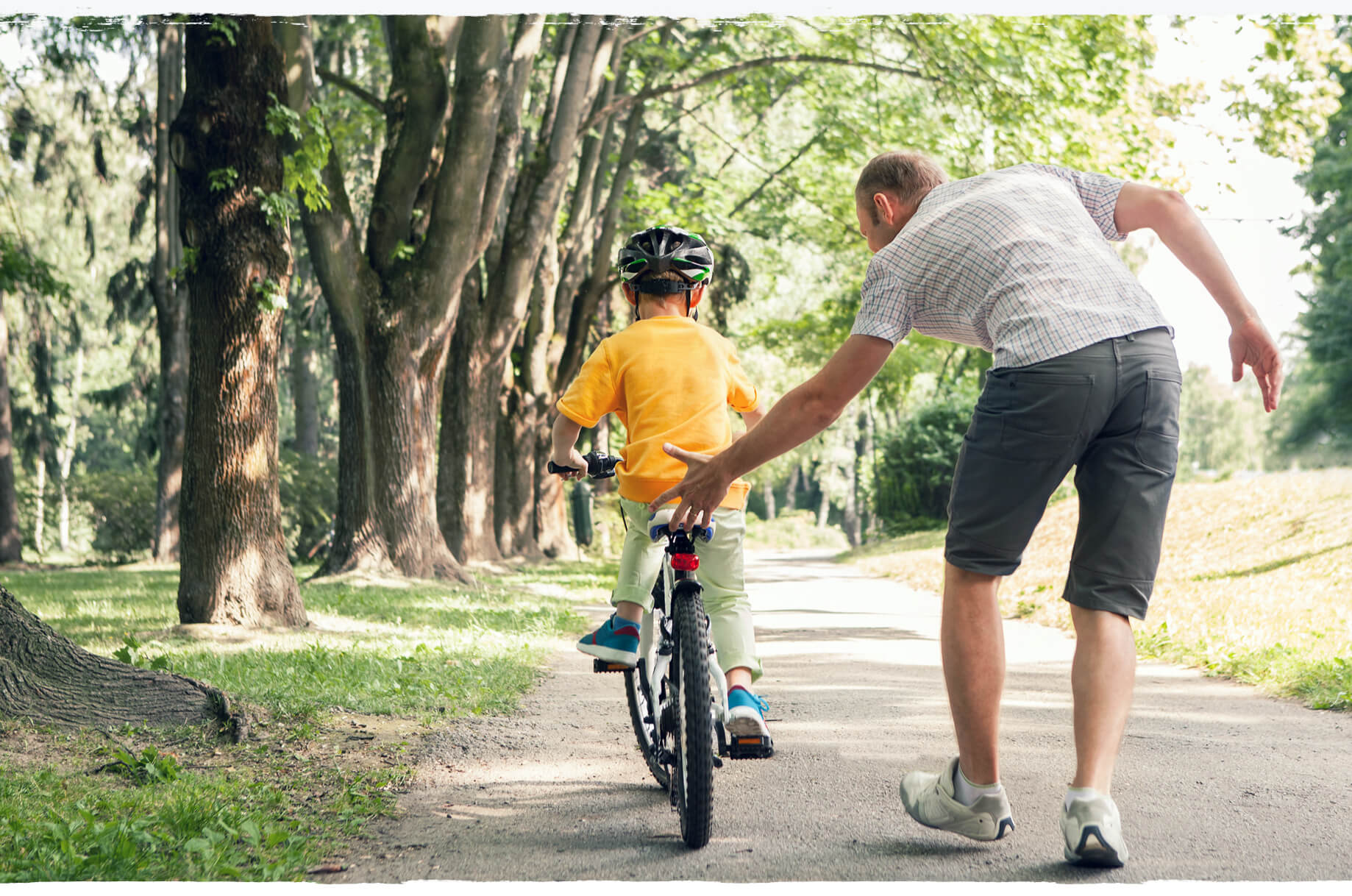 A man teaching a boy to ride a bike on a tree-lined park path